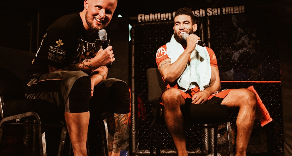 Two individuals are seated on a stage, each holding a microphone and smiling. They are in a sports-related setting with one wearing a casual black outfit and the other in sportswear, with a towel around his neck. The background includes elements of a fighting or martial arts cage.