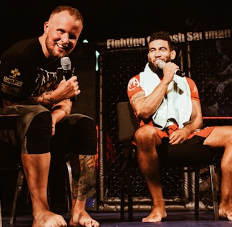 Two individuals are seated on a stage, each holding a microphone and smiling. They are in a sports-related setting with one wearing a casual black outfit and the other in sportswear, with a towel around his neck. The background includes elements of a fighting or martial arts cage.