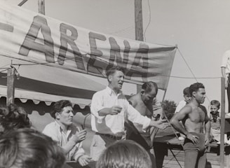 A group of men, some shirtless, are gathered around a platform or stage. A man in a white shirt appears to be speaking or gesturing to a crowd. A large sign with the word 'ARENA' is prominently displayed in the background. The scene seems to be set outdoors on a sunny day.
