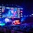 A large esports arena featuring a massive screen displaying a video game, surrounded by sponsor logos and team names. The auditorium is filled with spectators watching the gaming event. Neon blue and purple lights illuminate the stage, creating a vibrant atmosphere.