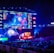 A large esports arena featuring a massive screen displaying a video game, surrounded by sponsor logos and team names. The auditorium is filled with spectators watching the gaming event. Neon blue and purple lights illuminate the stage, creating a vibrant atmosphere.