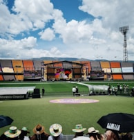 A sports stadium with a large stage set up on the field, hosting an outdoor event. Spectators wearing colorful hats sit in the foreground. The seating areas display vibrant colors, and a clear blue sky with scattered clouds is visible. Large screens display graphics related to the event.
