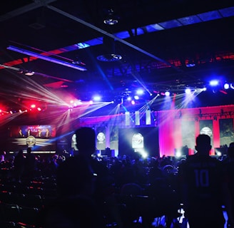 A dimly lit indoor arena filled with an audience. Bright stage lights in red and blue illuminate multiple large screens displaying graphics.