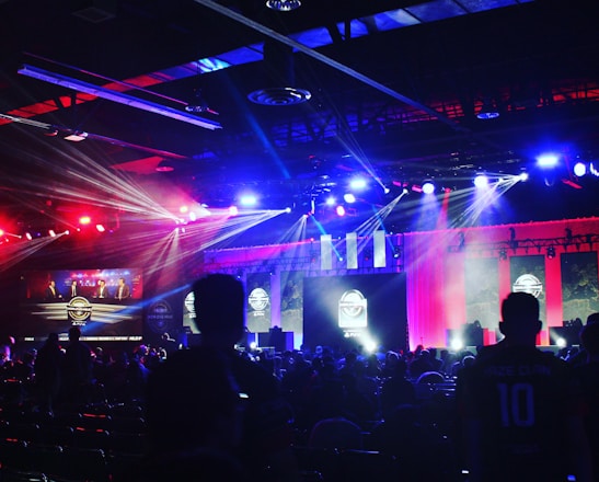 A dimly lit indoor arena filled with an audience. Bright stage lights in red and blue illuminate multiple large screens displaying graphics.