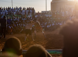 A vibrant wrestling match taking place outdoors, surrounded by a large crowd of spectators. The atmosphere is charged with excitement as people stand and sit around the dirt arena, intently watching the competitors. The sun is setting, casting a warm glow over the scene.