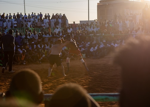 A vibrant wrestling match taking place outdoors, surrounded by a large crowd of spectators. The atmosphere is charged with excitement as people stand and sit around the dirt arena, intently watching the competitors. The sun is setting, casting a warm glow over the scene.