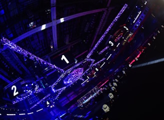 A large indoor arena with colorful and vibrant lighting, featuring a center stage surrounded by rigging and structures. Large numbered screens are visible, indicating marked sections from one to five. Various pieces of equipment and people are scattered around the venue.