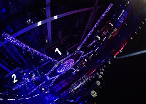 A large indoor arena with colorful and vibrant lighting, featuring a center stage surrounded by rigging and structures. Large numbered screens are visible, indicating marked sections from one to five. Various pieces of equipment and people are scattered around the venue.