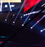 An array of bright stage lights beams across a dark arena, creating a dynamic atmosphere with a large screen displaying various logos and text. A camera crane is visible in the foreground, suggesting a professional event setup.