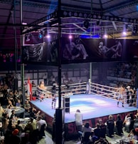 A crowded indoor arena with a boxing ring at the center, surrounded by an audience seated at tables. The scene is illuminated by overhead lights, casting a vibrant glow over the ring where two fighters are preparing for a match. Spectators are engaged, capturing the event with cameras and enjoying the lively atmosphere.