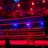 An empty boxing ring is illuminated by dramatic red and blue spotlights inside a dimly-lit arena. A small stool is placed in the center of the ring, surrounded by ropes. The arena is adorned with banners depicting action scenes, and a few silhouettes of spectators can be seen in the background.