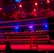 An empty boxing ring is illuminated by dramatic red and blue spotlights inside a dimly-lit arena. A small stool is placed in the center of the ring, surrounded by ropes. The arena is adorned with banners depicting action scenes, and a few silhouettes of spectators can be seen in the background.