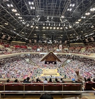 A large indoor arena packed with spectators seated in tiers around a central platform. The platform features traditional Japanese decor, indicating it may be used for sumo wrestling. The atmosphere is bustling, with people filling the seats and many spectators visible in the foreground.