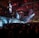 A large indoor event with a stage featuring bright screens and spotlights, attended by a substantial crowd. The venue has a high ceiling with industrial beams and red lighting accents. The atmosphere resembles a concert or a conference with a focus on visual effects.