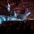 A large indoor event with a stage featuring bright screens and spotlights, attended by a substantial crowd. The venue has a high ceiling with industrial beams and red lighting accents. The atmosphere resembles a concert or a conference with a focus on visual effects.