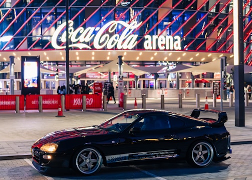 A sleek black sports car is parked in front of an illuminated arena entrance, with a large sign bearing the name of a famous beverage brand. The building features a modern, geometric facade with bright red and blue lights, creating a vibrant and lively atmosphere.